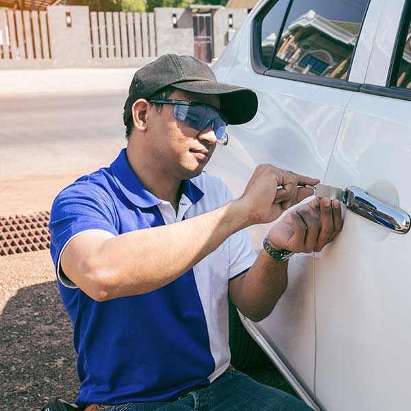 Man in blue shirt and cap using a lockpick to work on a white car door.