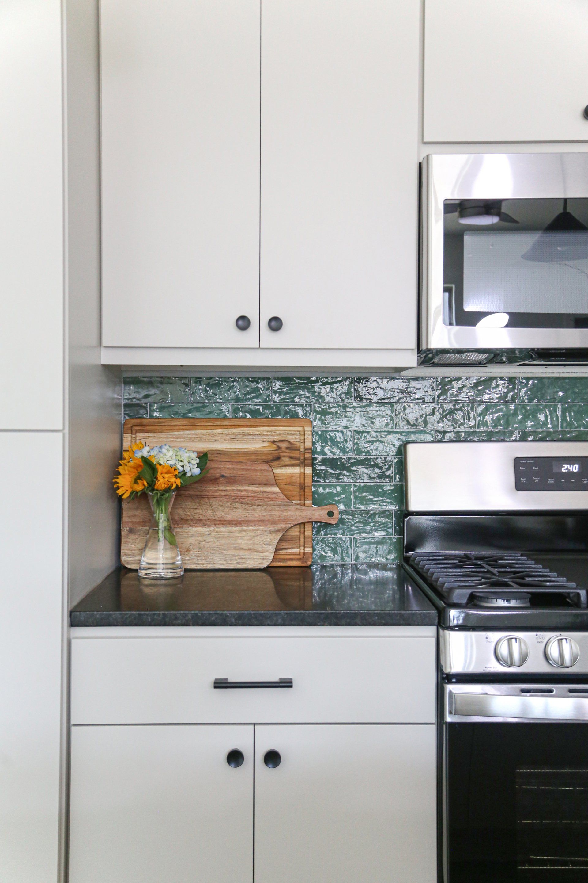 A kitchen with white cabinets , a stove , a microwave and a cutting board on the counter.