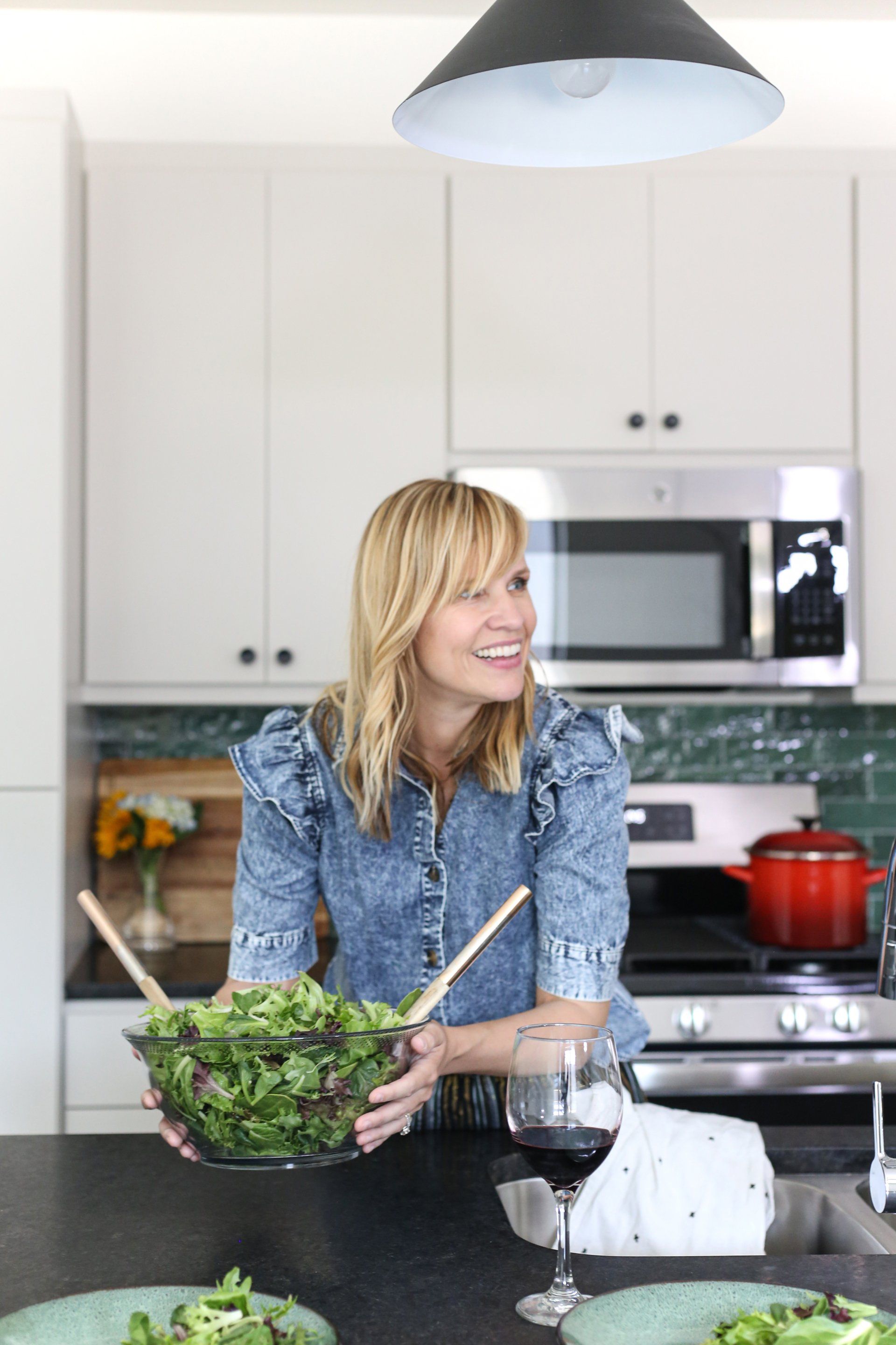 A woman is standing in a kitchen holding a bowl of salad and a glass of wine.