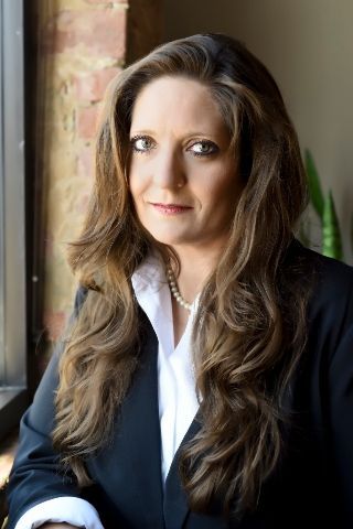 A woman in a black jacket and white shirt is standing in front of a window.