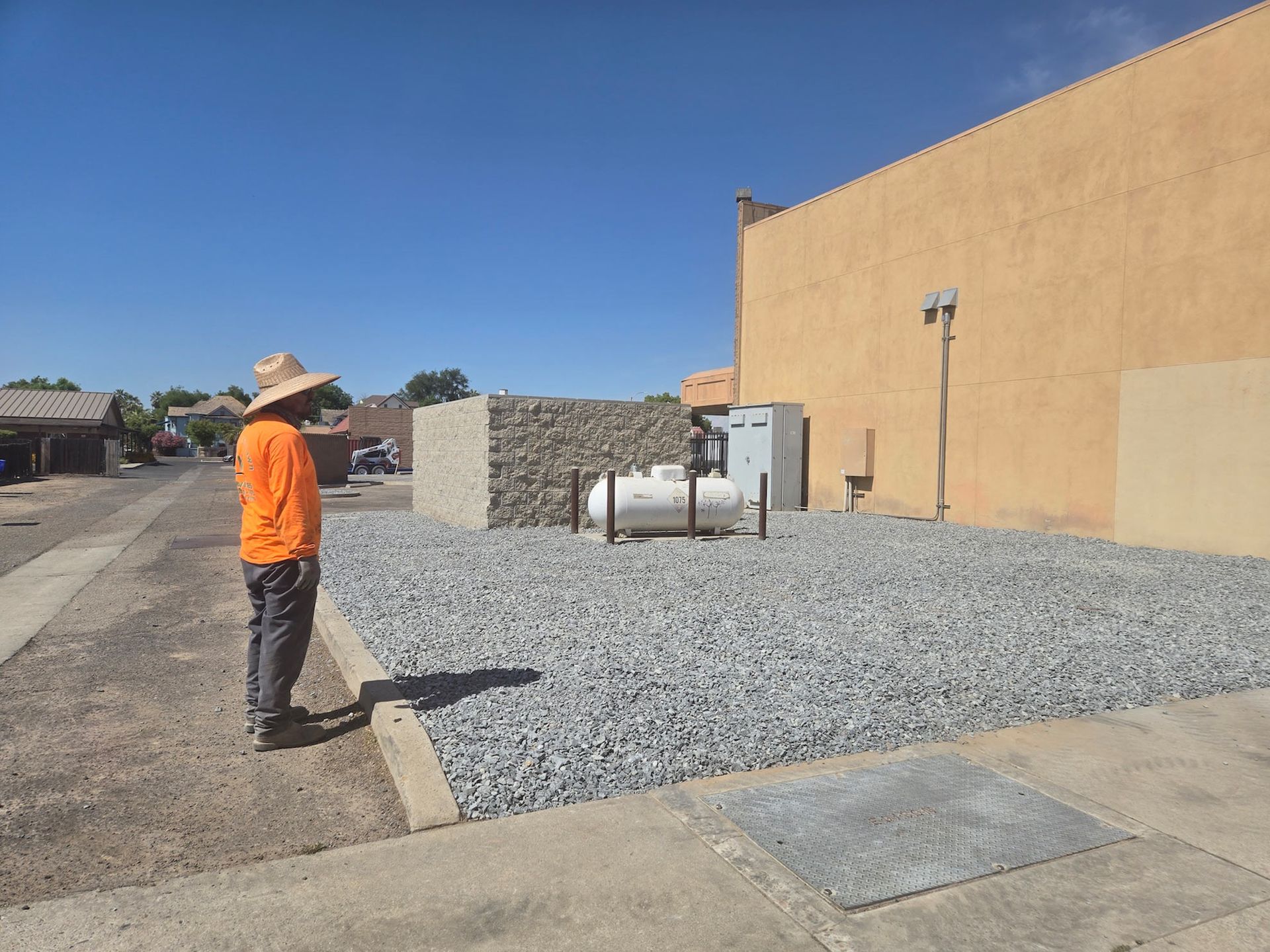 A person wearing an orange shirt and sun hat stands on a gravel patch viewing a small propane tank by a tan building.