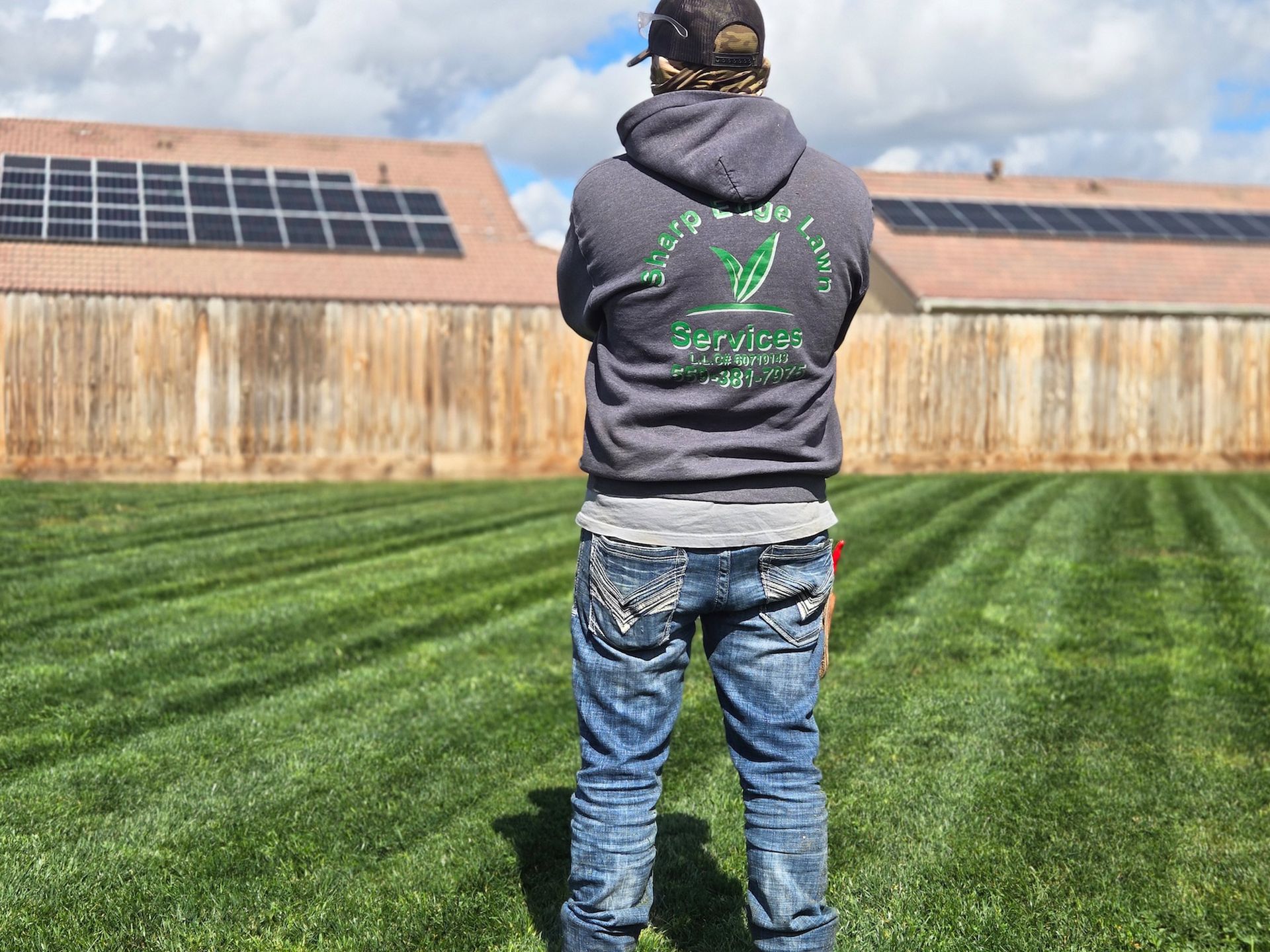 A person wearing a gray hoodie stands in a freshly mowed lawn, facing a wooden fence with houses and solar panels behind.
