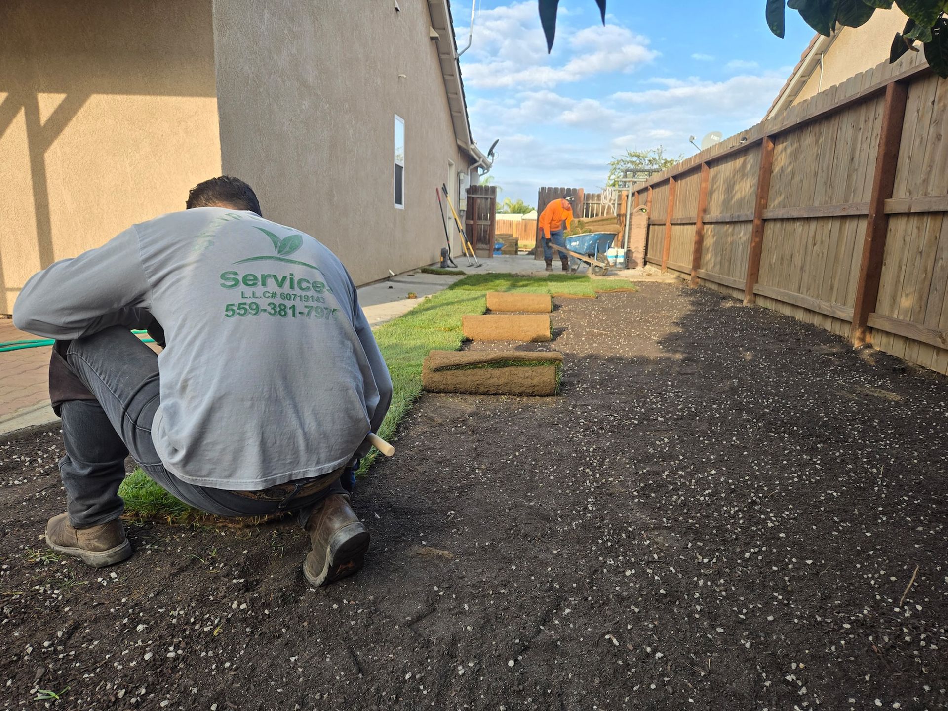 Landscapers install sod in a residential yard beside a wooden fence and a house.