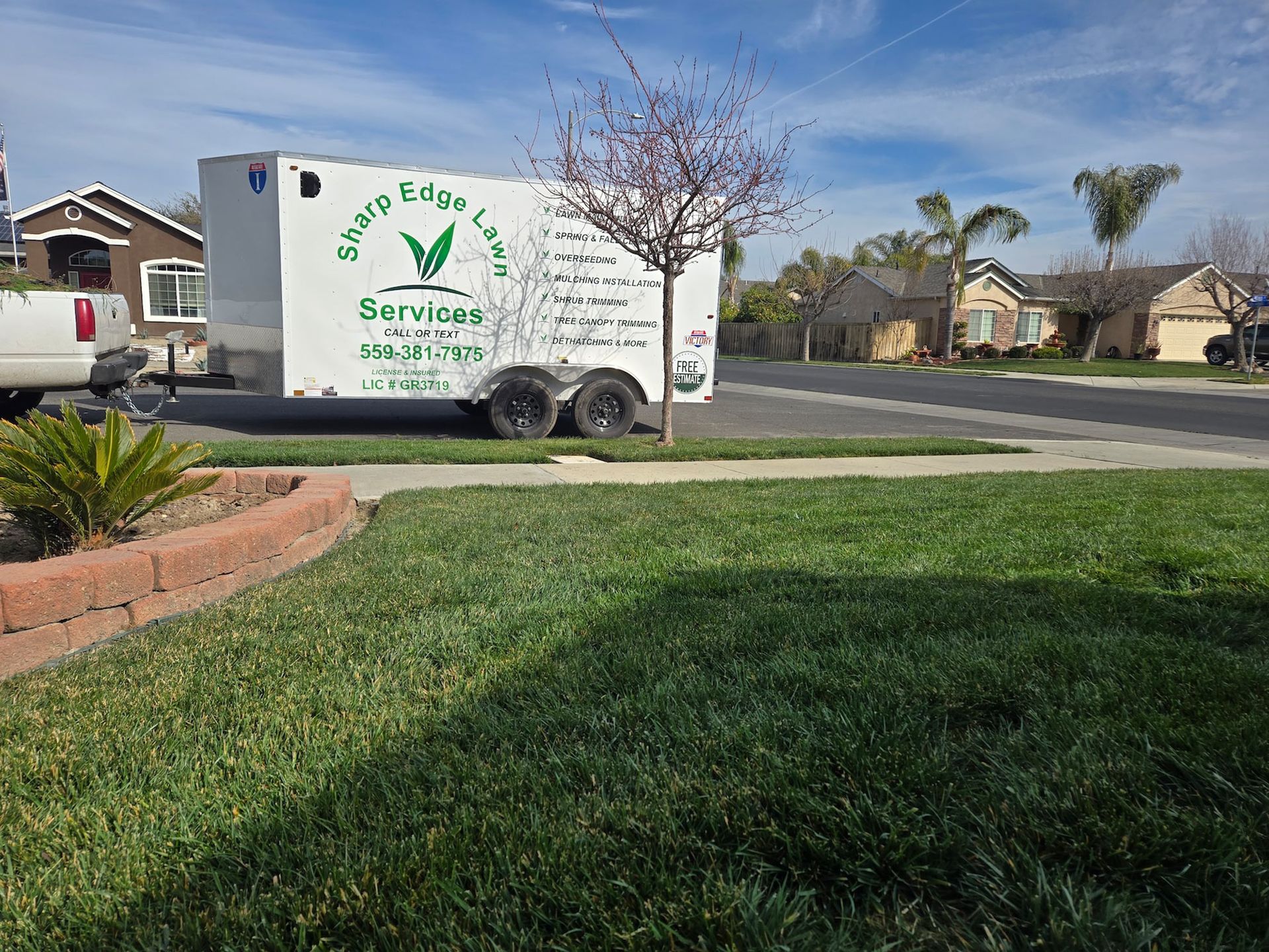 A white trailer parked on a suburban street, marked with the business name Sharp Edge Lawn Services and contact information.