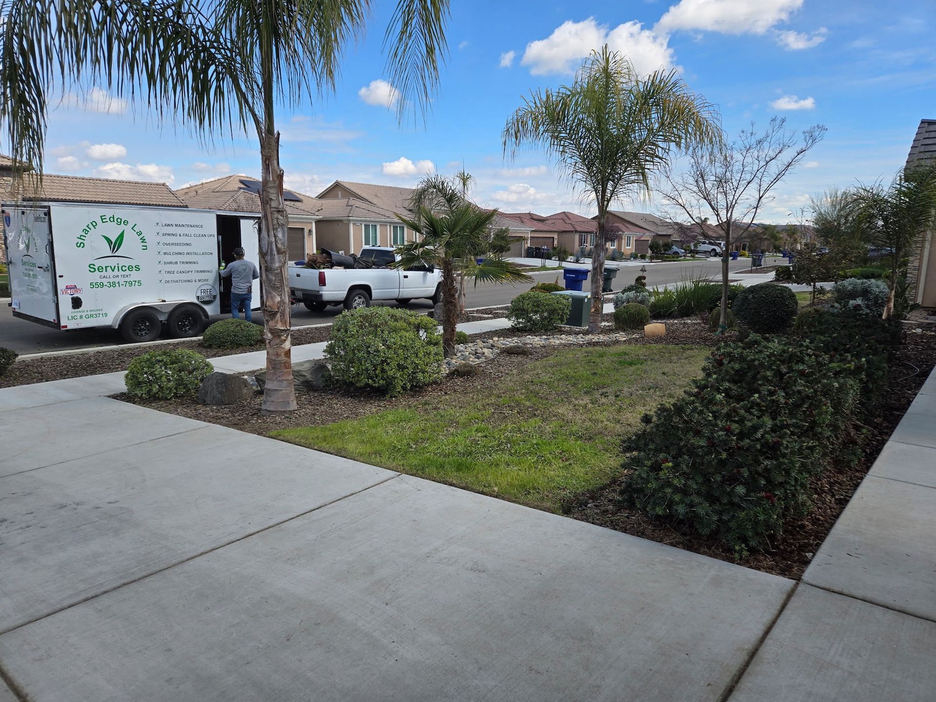 A white service trailer parked in a sunny residential neighborhood next to a landscaped front yard with palm trees.