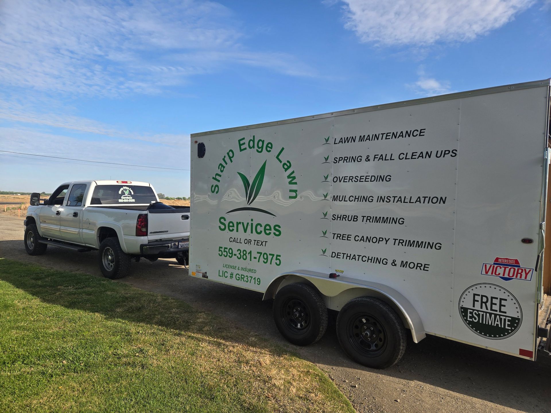 White pickup truck pulling a trailer for Sharp Edge Lawn Services, parked on a dirt path under a clear blue sky.