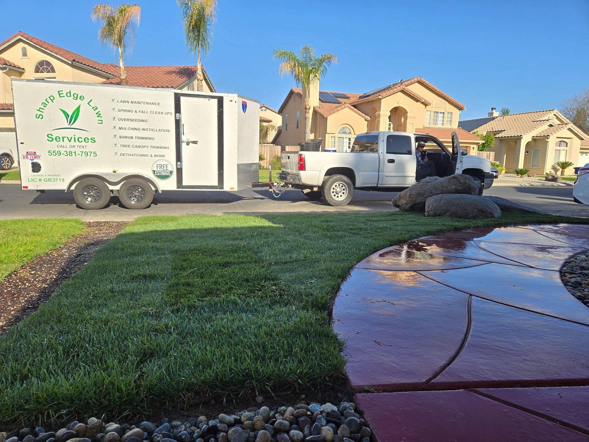 A white landscaping service truck and trailer parked on a suburban street next to a lush green lawn and paved path.