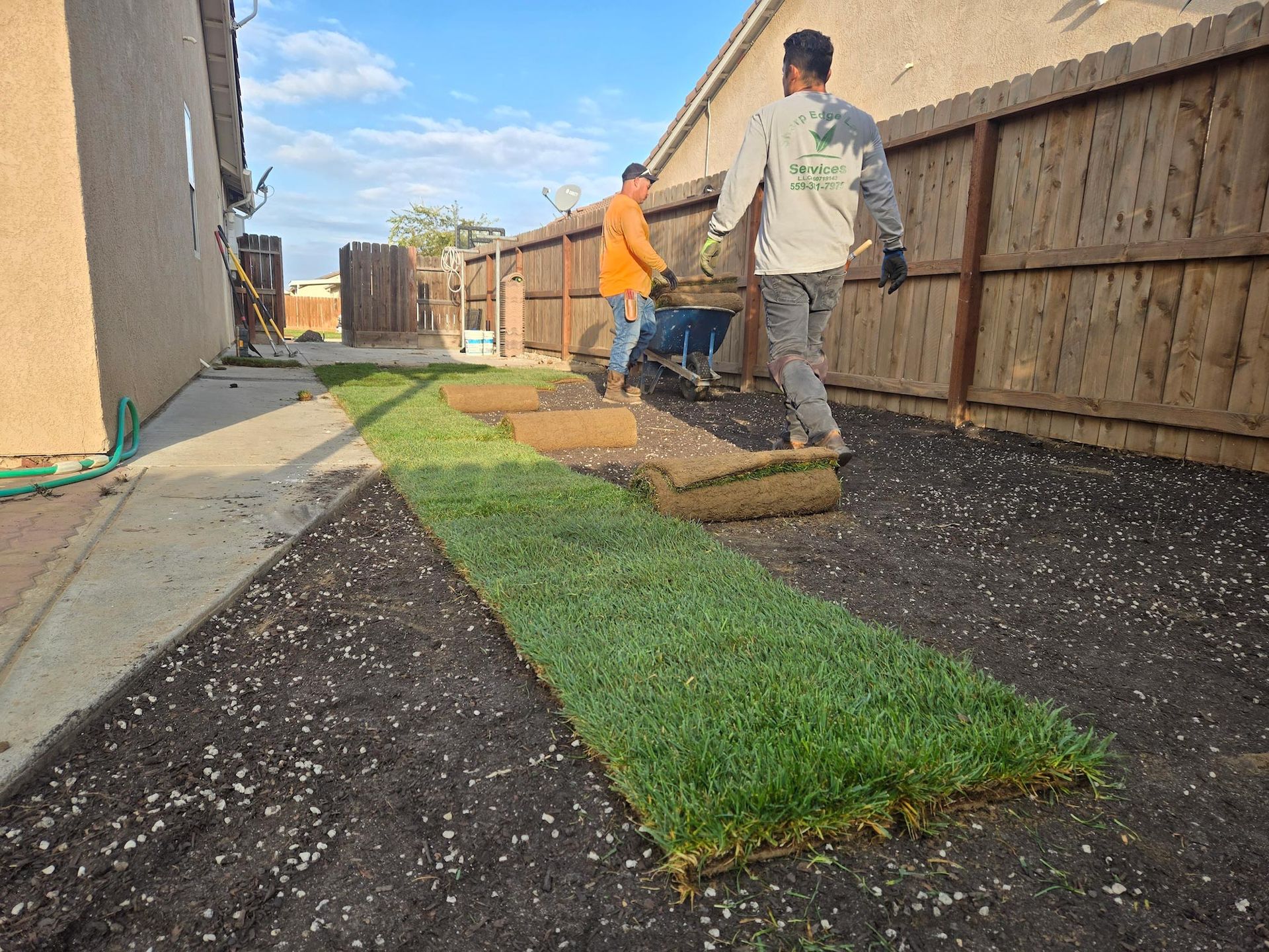 Two workers install rolls of green sod on dark soil in a residential backyard next to a wooden fence and house wall.