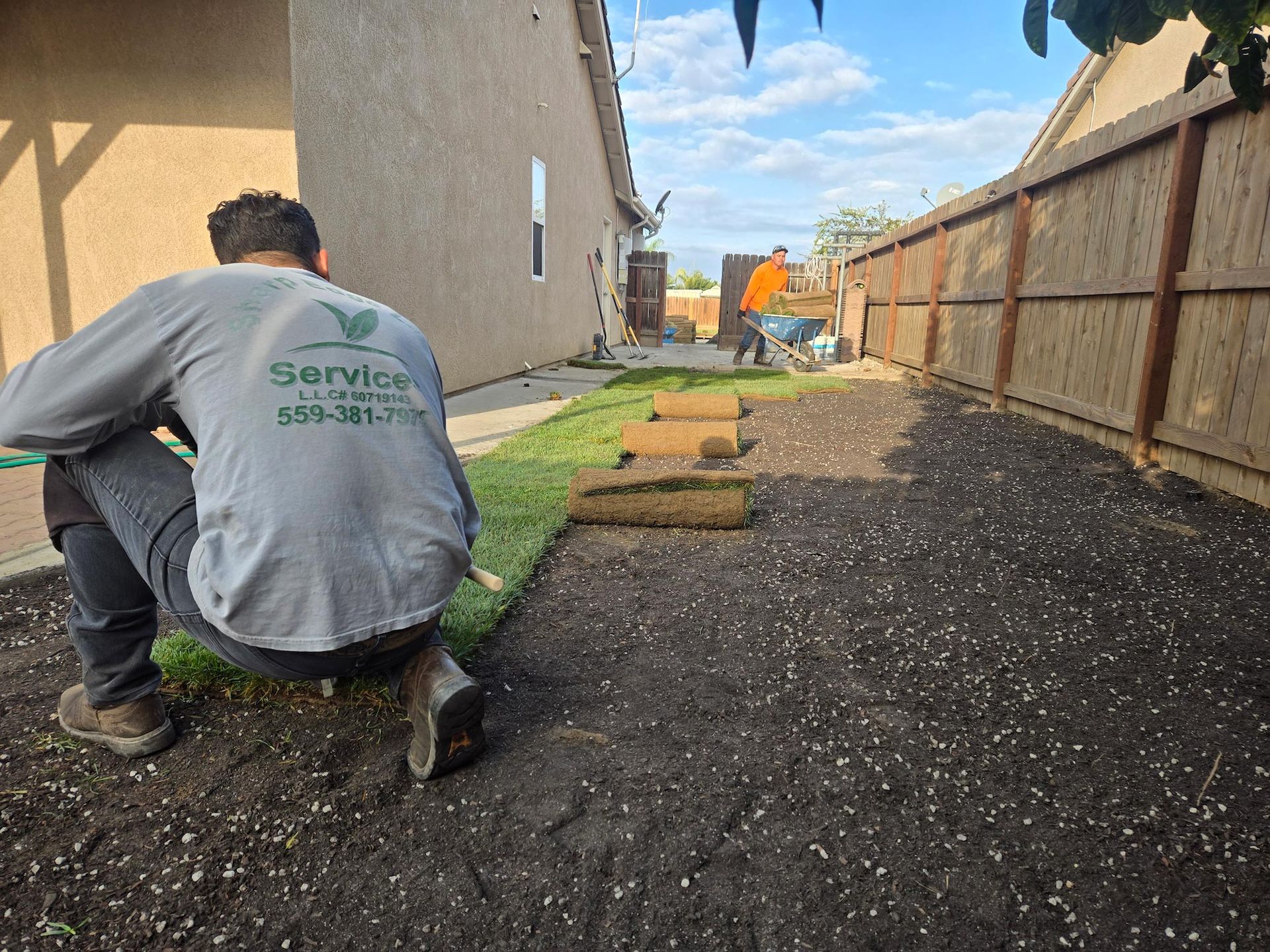 Two workers laying sod in a backyard next to a residential house and a wooden fence.