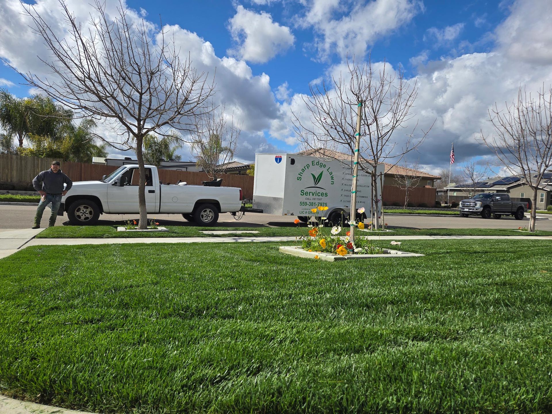 A person stands next to a white pickup truck pulling a utility trailer on a suburban street with green lawns and trees.