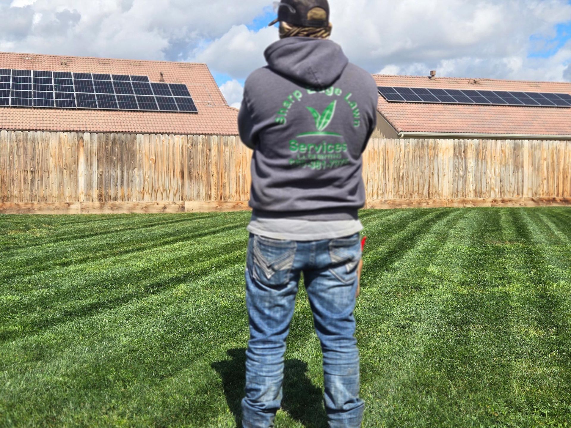 A person stands in a backyard with a neatly mown lawn, facing a wood fence and houses with solar panels on the roof.