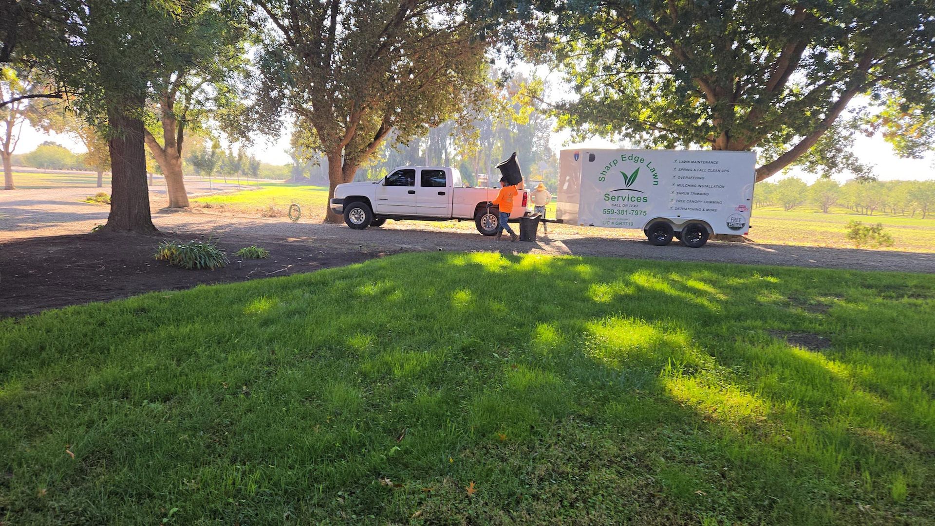 A white pickup truck pulling a trailer with a logo is parked under trees next to a green lawn.
