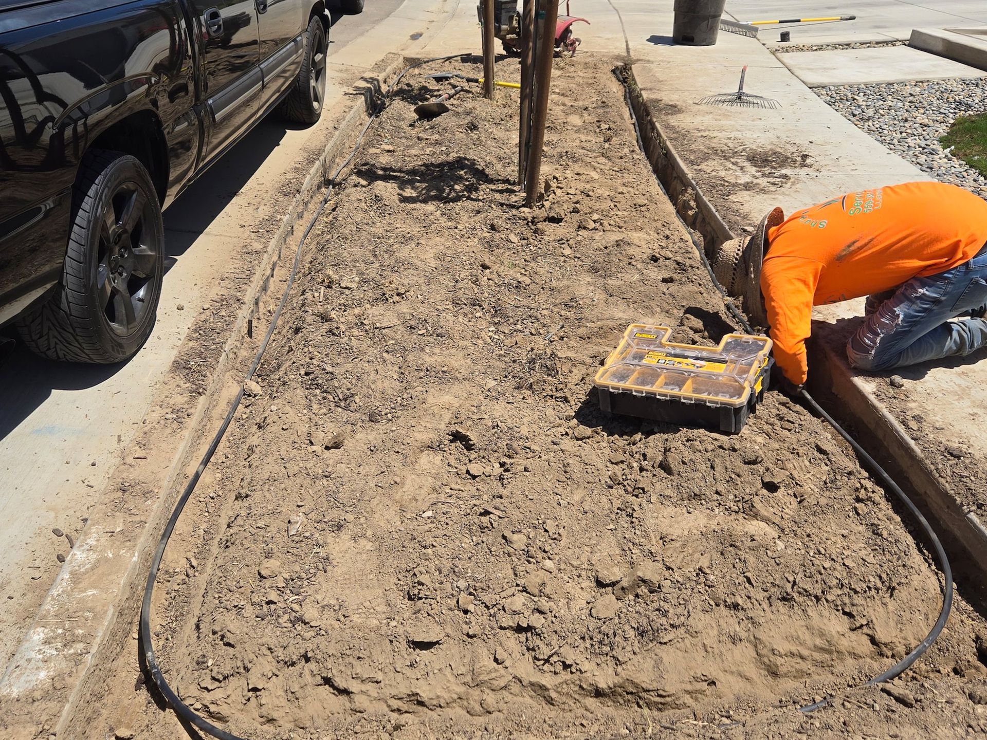 A worker in an orange shirt kneels by a trench, installing irrigation tubing in a dirt flowerbed next to a parked truck.