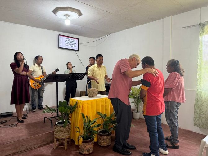 People praying in a small chapel, with musicians at the front and one person kneeling in prayer.