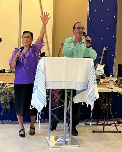 Two people speaking onstage, one raising a hand, behind a white podium at an indoor event
