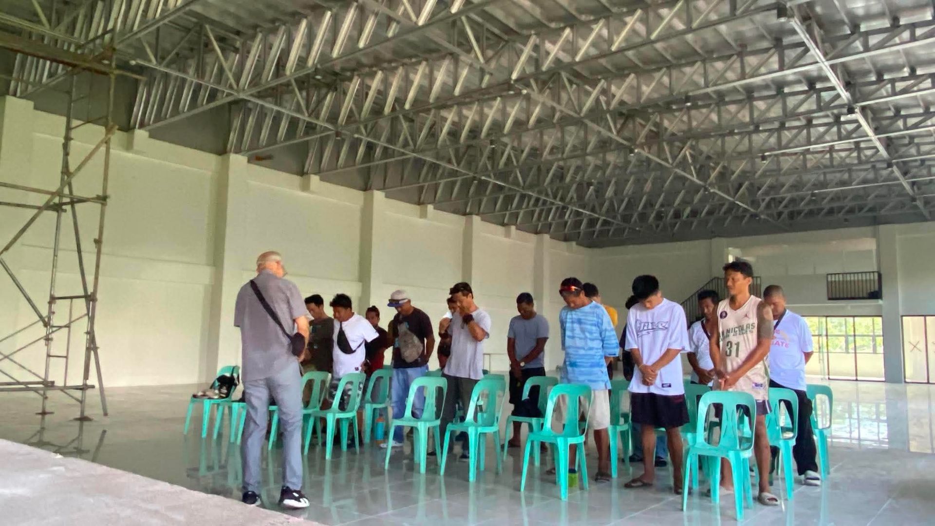 People gathered around teal chairs in a large indoor hall with a metal roof.