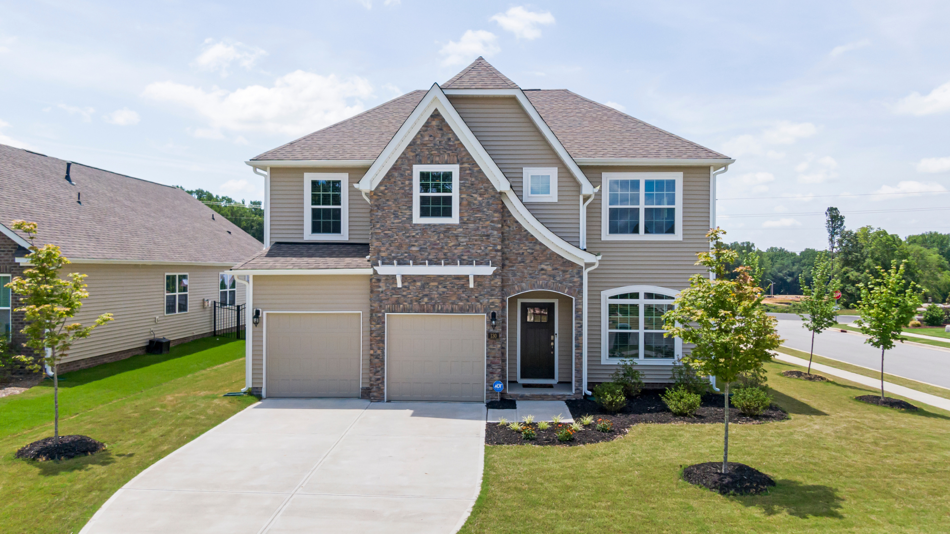 Two-story beige house with brick accents, two-car garage, and manicured lawn on a sunny day.