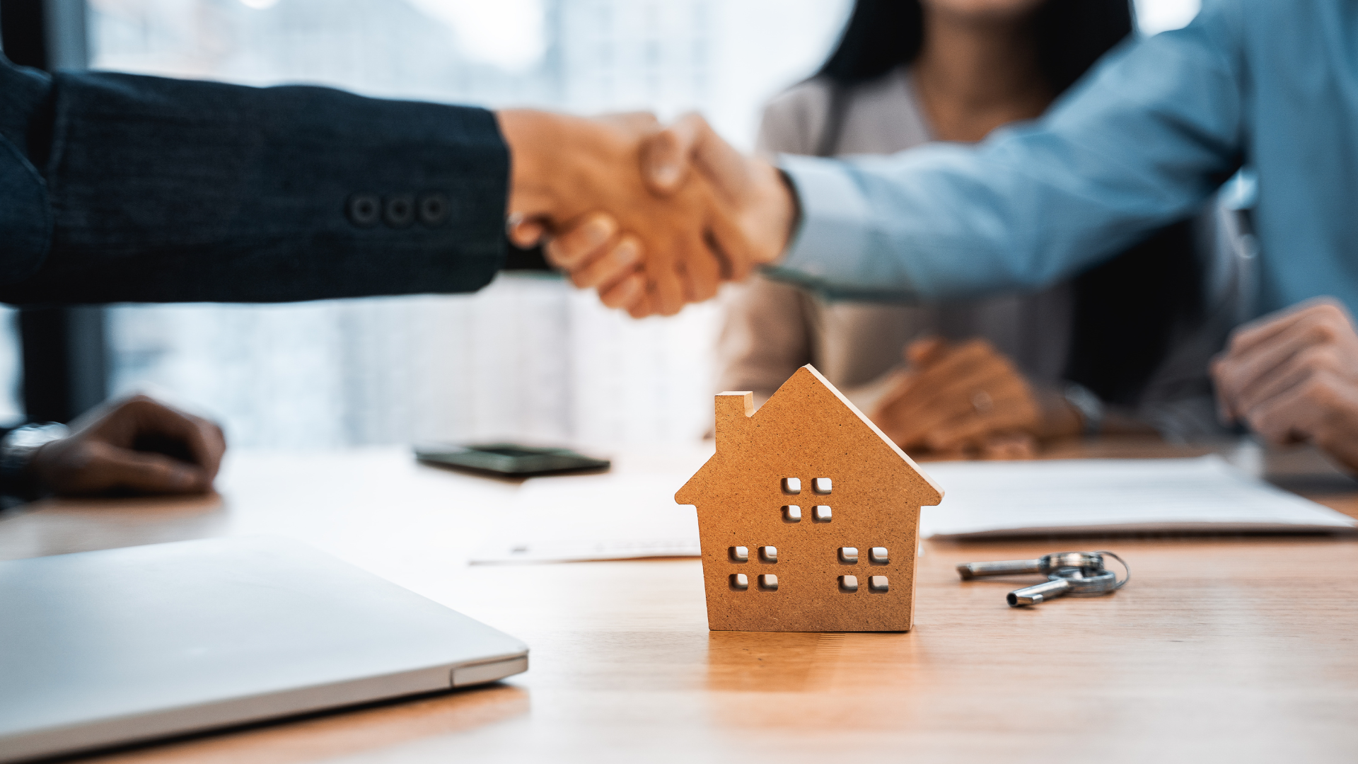 Handshake sealing a real estate deal; wooden house model, keys, and documents on a table.