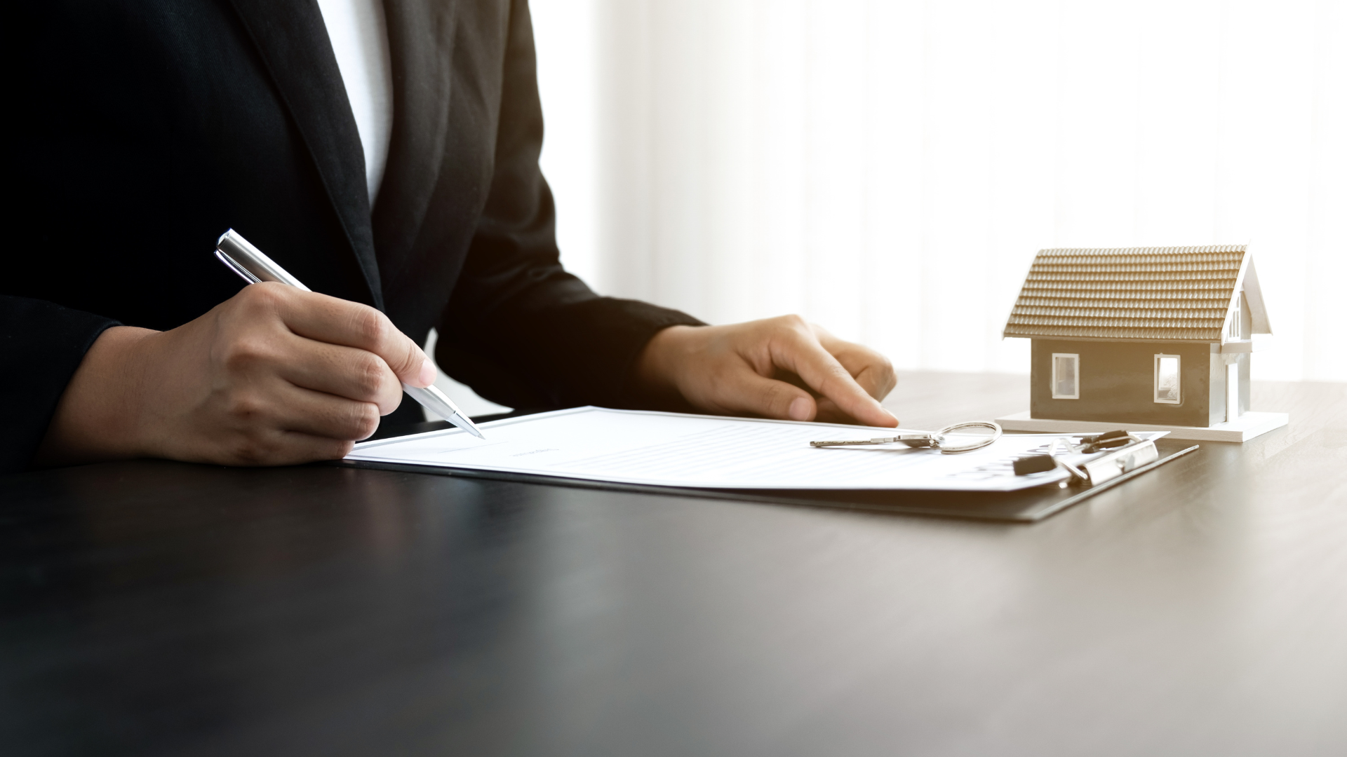 Person in a blazer signs a document on a clipboard next to a small house model.