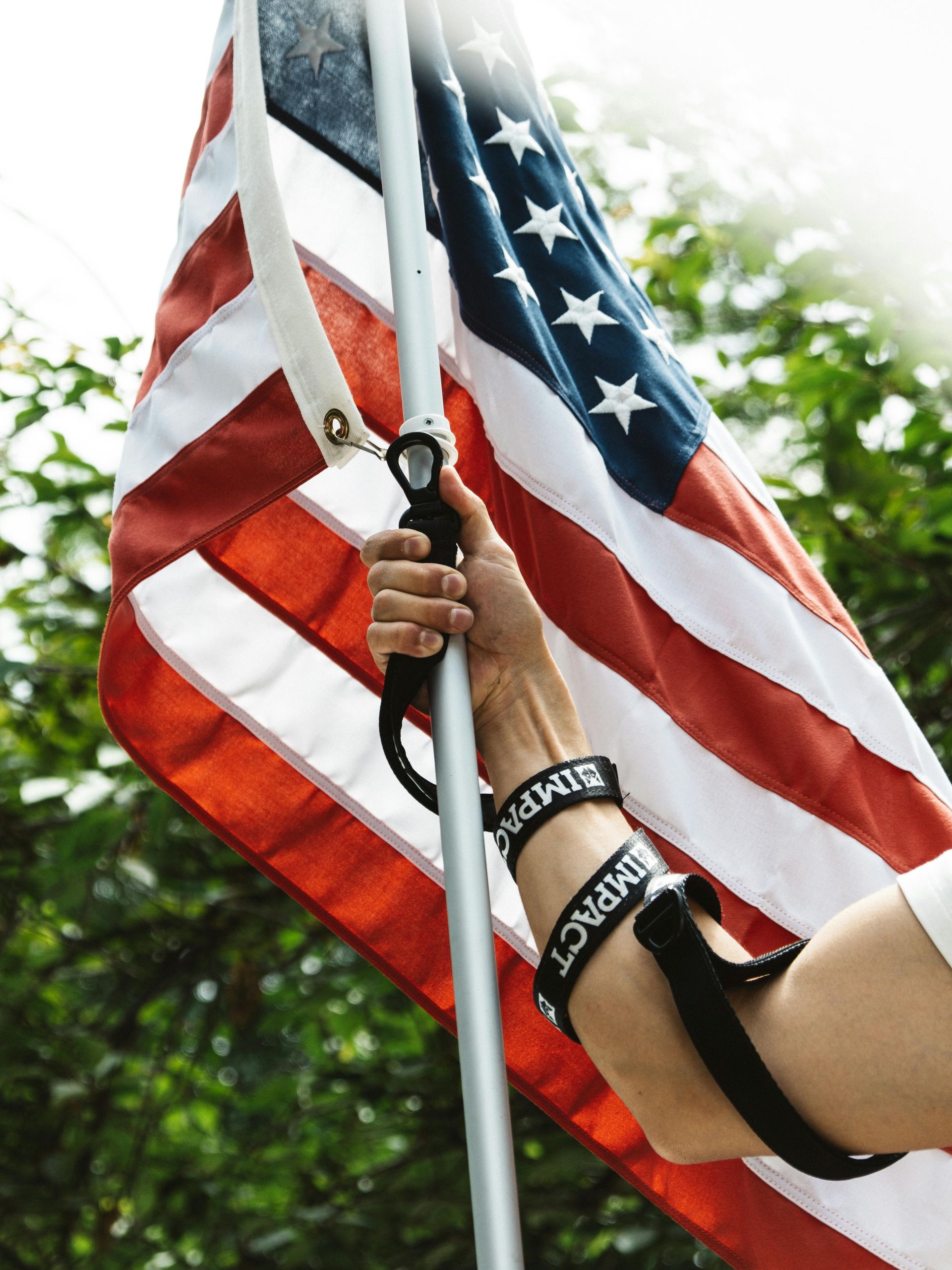 Hand holding American flag, close-up. Arm with black wristbands against a blurred green backdrop in Post Falls Idaho.