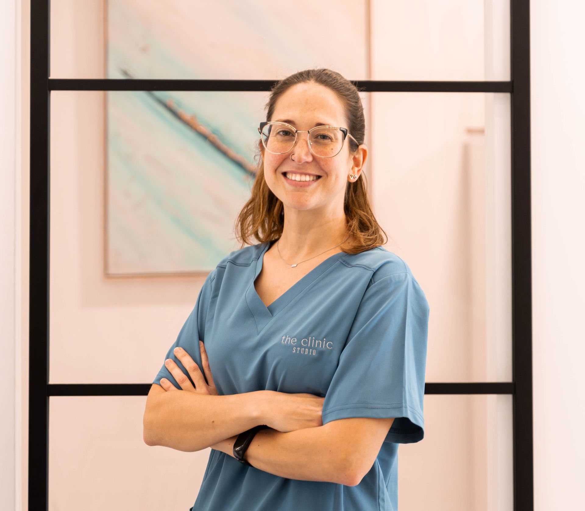 Una mujer con una blusa azul y gafas está de pie con los brazos cruzados y sonriendo.