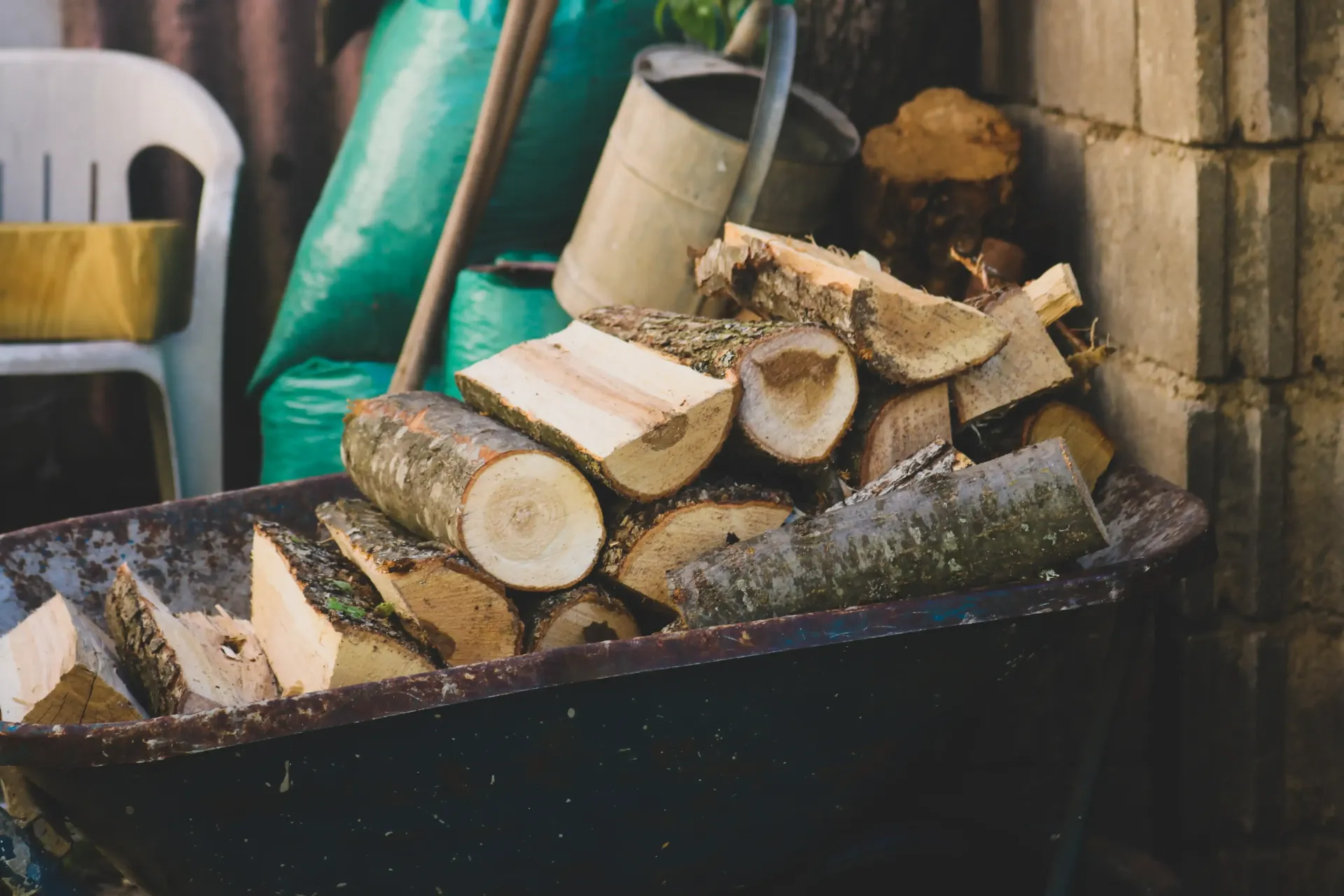 Freshly cut logs stacked in a black wheelbarrow beside a white chair and garden wall