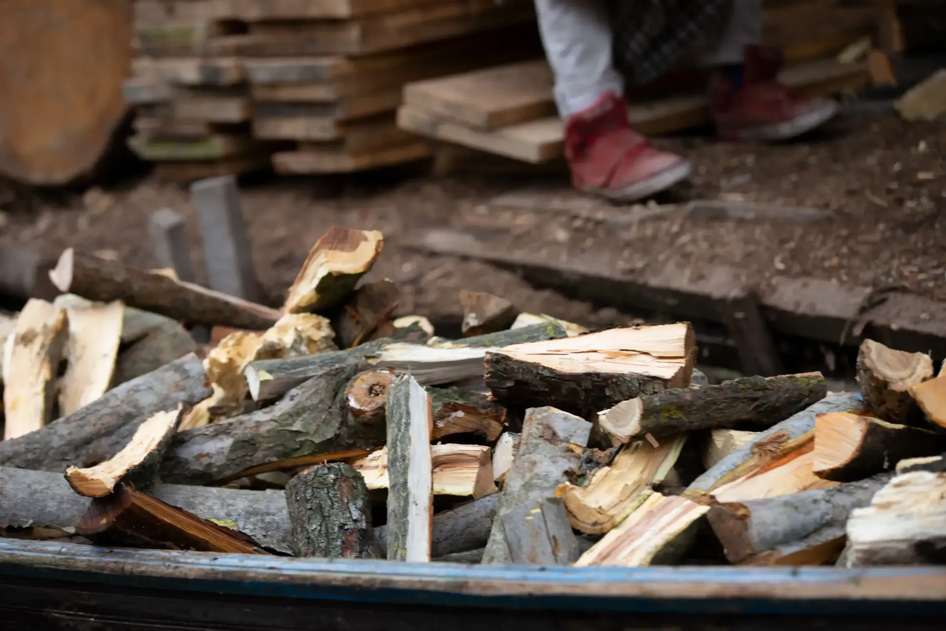 Split firewood piled outdoors near a person's boots and stacked lumber.