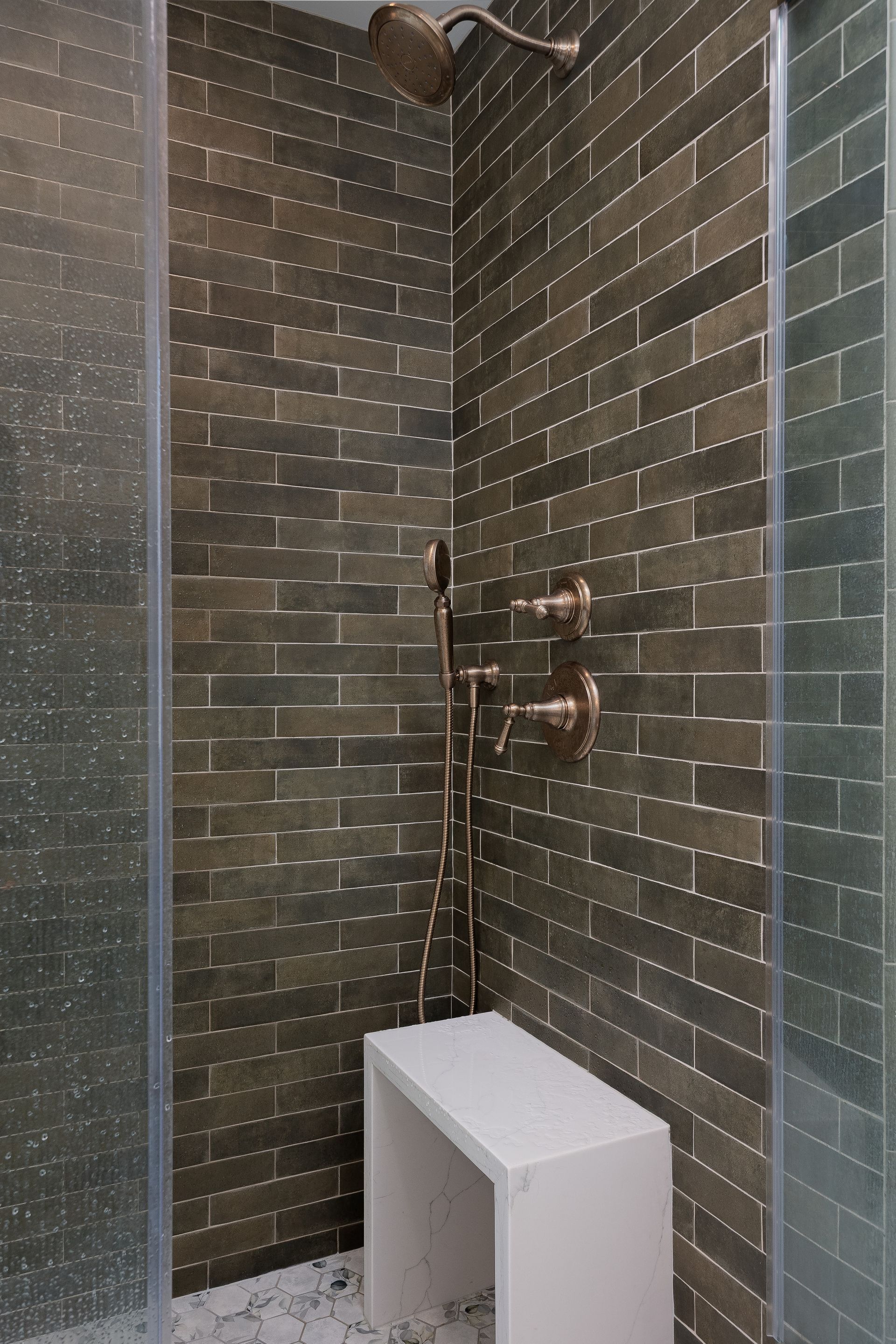 Shower with green-brown brick-like tile, brushed gold fixtures, a white bench, and a glass door.
