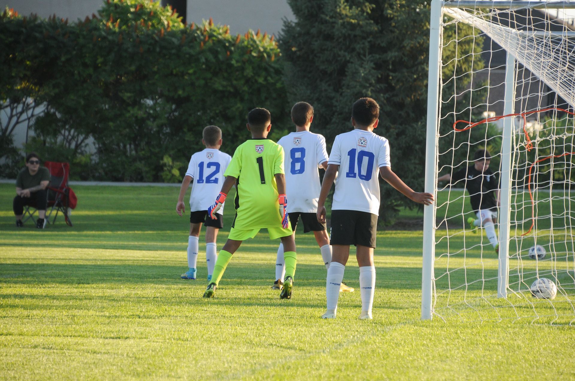 Soccer players in white and blue jerseys near a goal; one in a green jersey.