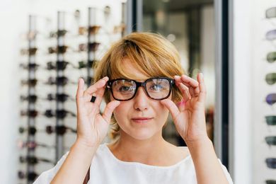 Woman in glasses looking at the camera in an eyewear store, holding the glasses with both hands.