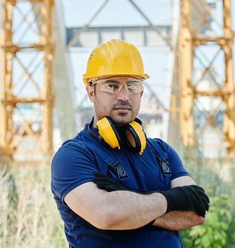 Construction worker with yellow hard hat, safety glasses, and ear protection, arms crossed on site.