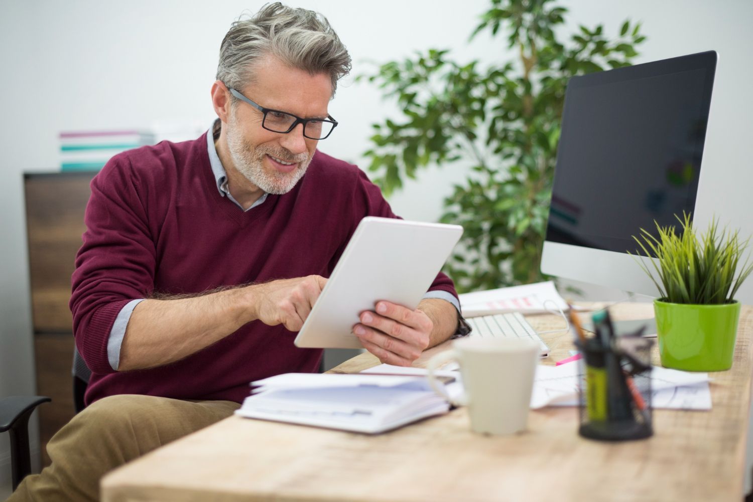 Man at a desk using a tablet beside a computer, papers, and a coffee cup in a bright office