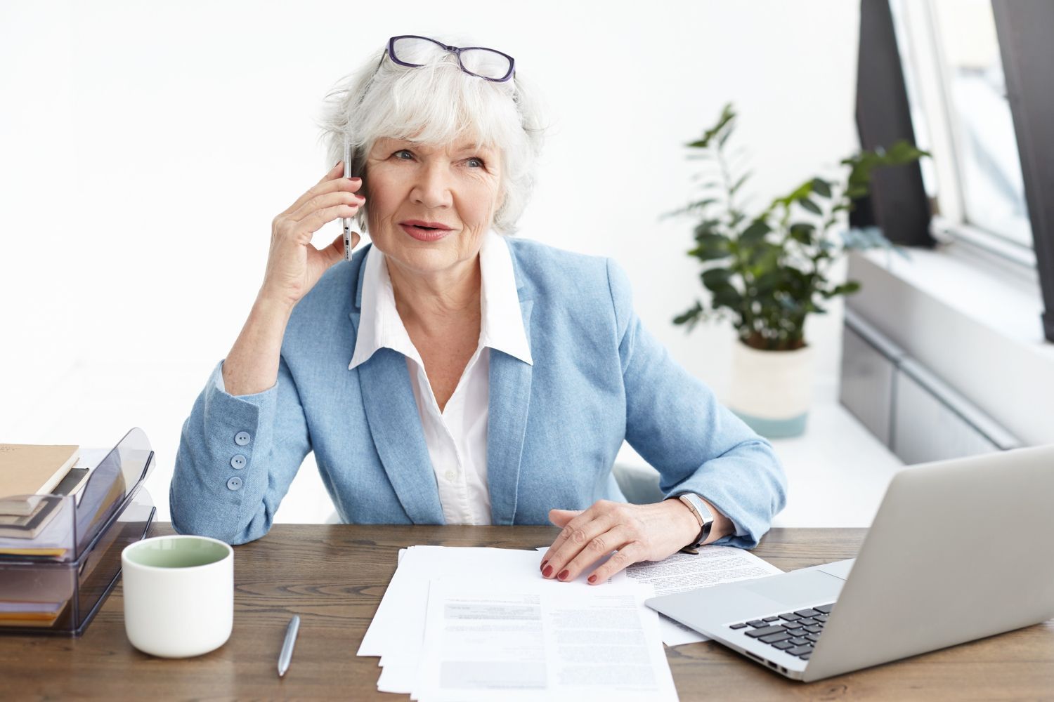 Woman at desk with laptop, papers, coffee mug, and plant, speaking on a phone in a bright office