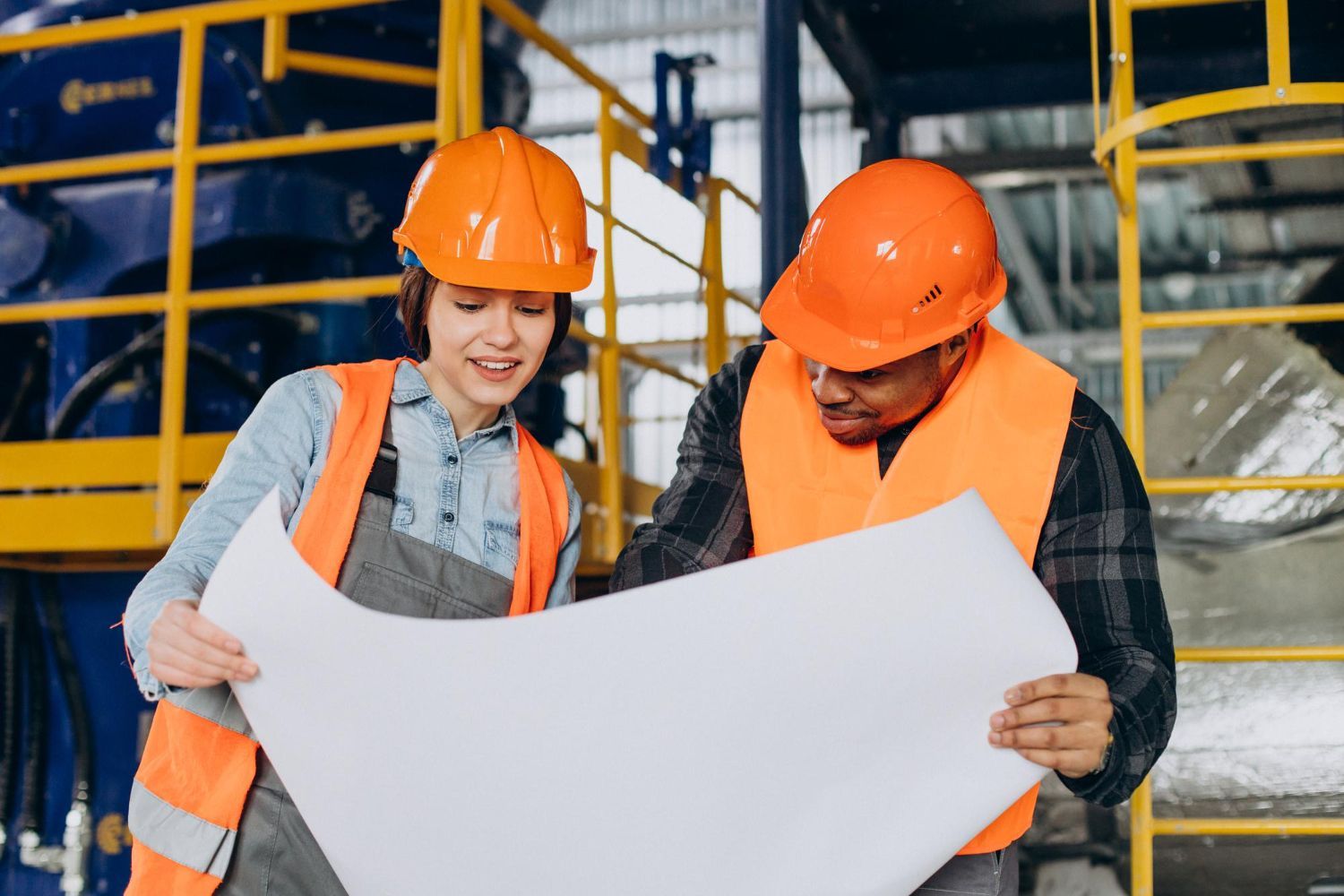 Two workers in orange hard hats reviewing a large blueprint in an industrial setting