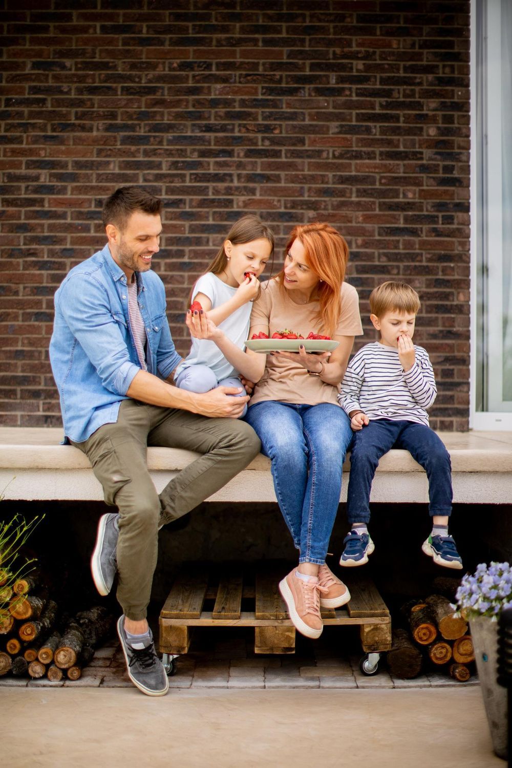 Family sitting on a porch step, sharing a snack and smiling against a brick wall.