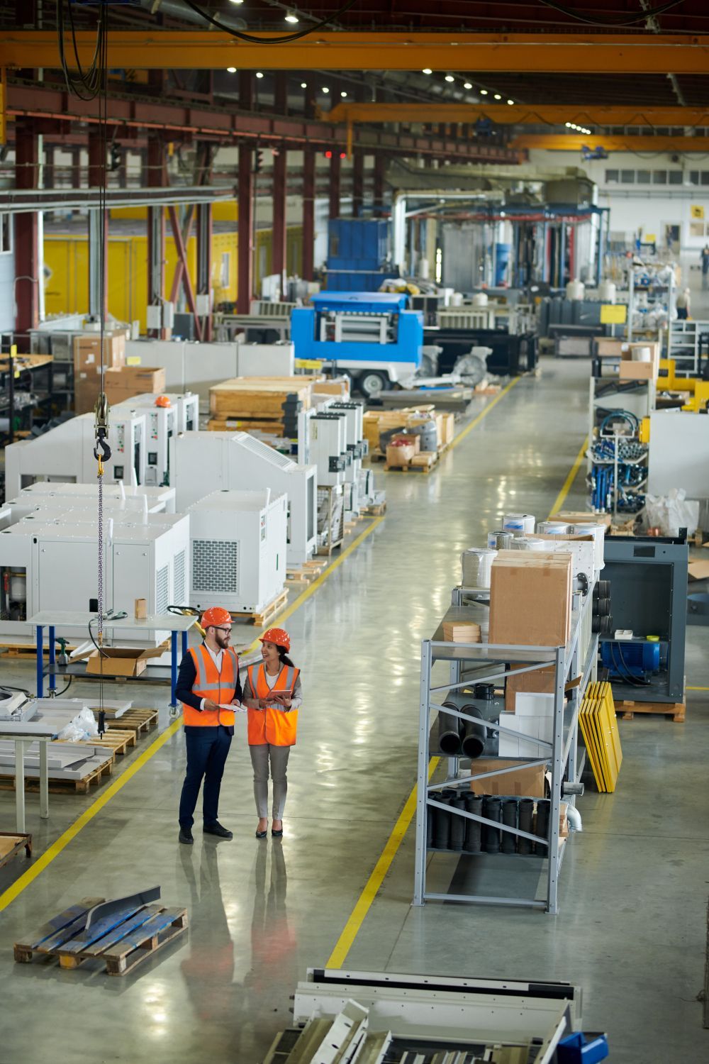Factory floor with two workers in orange vests beside large industrial machinery and equipment pallets