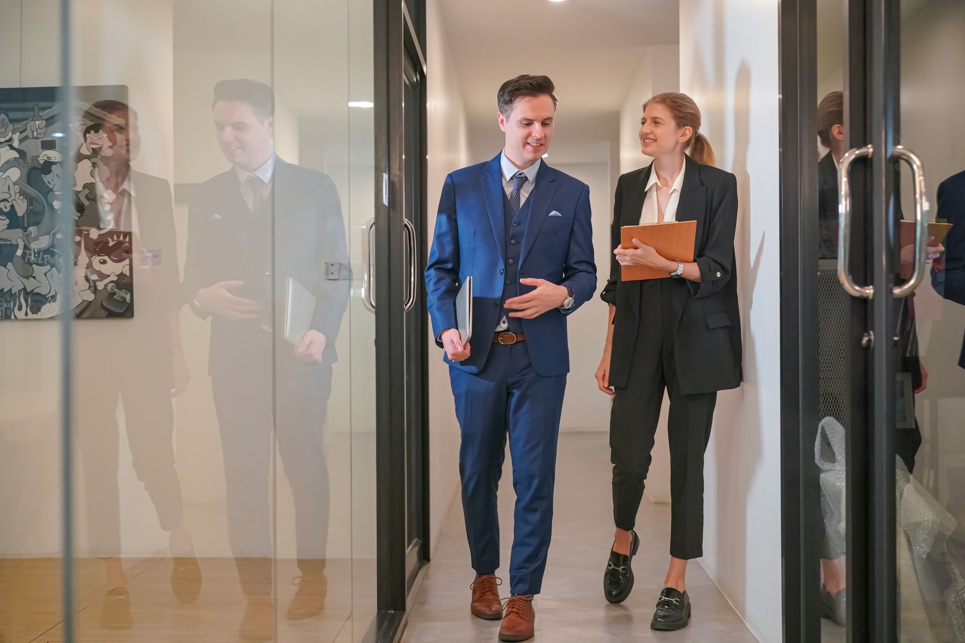Two professionals walking through an office hallway, one in a blue suit and one in black holding a folder.