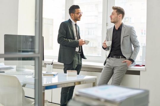 Two coworkers in suits talking by a bright office window, one holding a coffee cup.