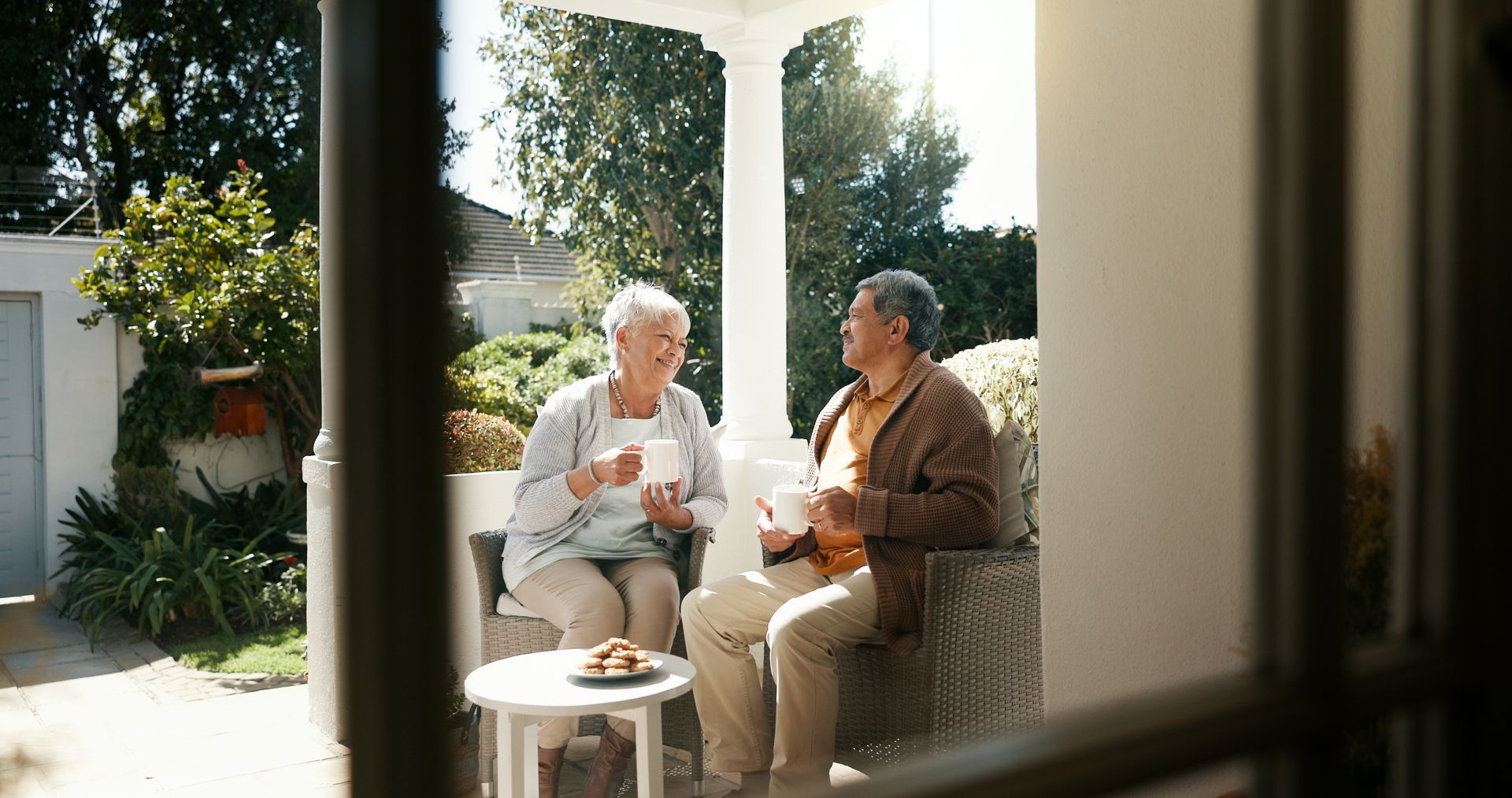 Two people chatting on a sunlit patio, seated beside a small table and potted plants.