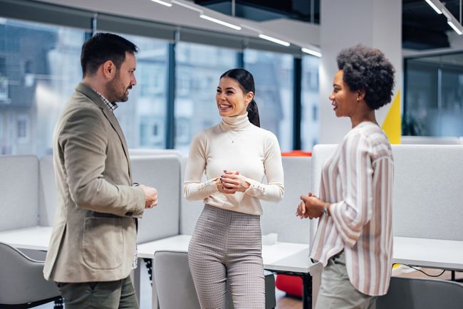 Three coworkers talking in a modern office, smiling and standing near desks.