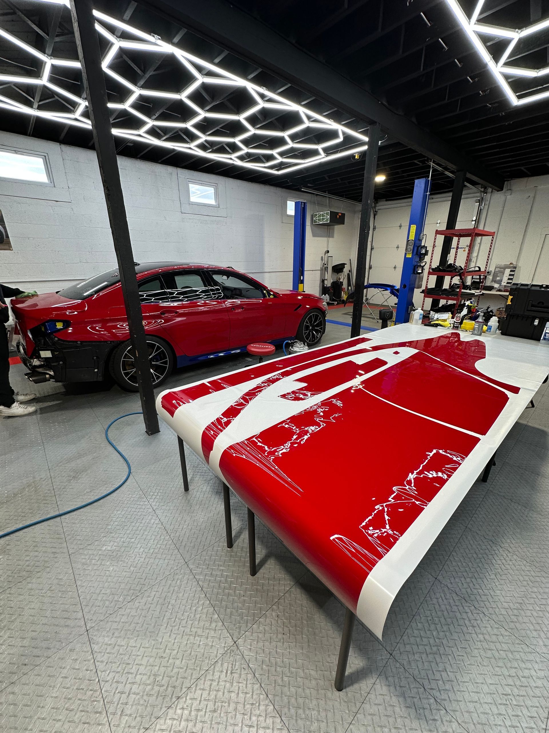 A red car sits in a professional auto shop, with a large piece of matching red vinyl wrap laid out on a table in front.