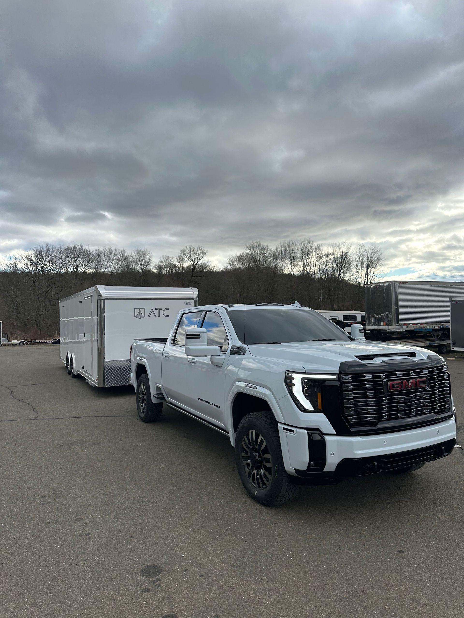 A white GMC truck towing a silver enclosed trailer parked on a paved lot under a cloudy sky.