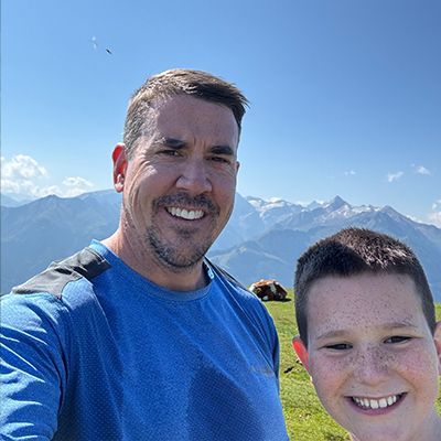 Two smiling people taking a selfie on a grassy mountain overlook under a clear blue sky.