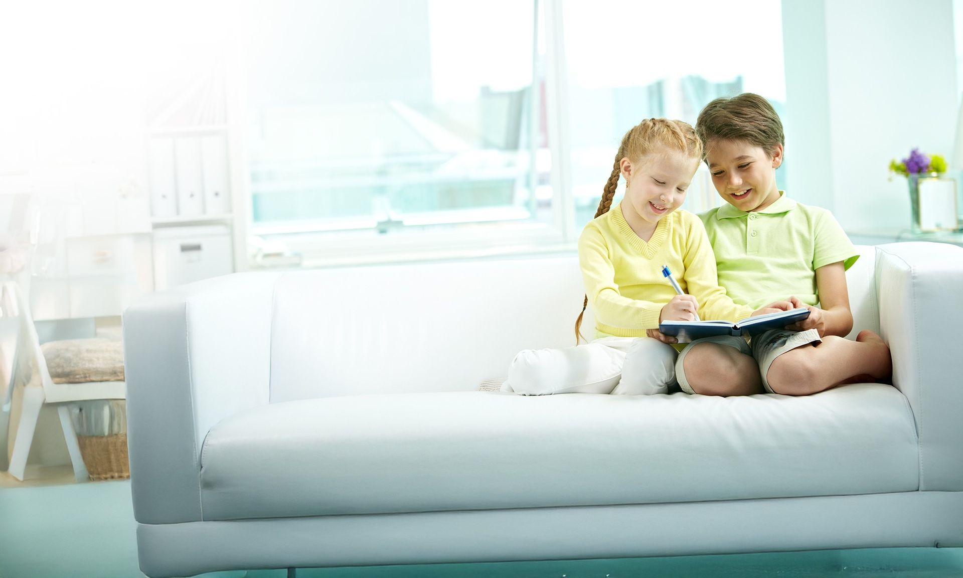 Two children sit on a white couch, looking at a book in a bright living room.