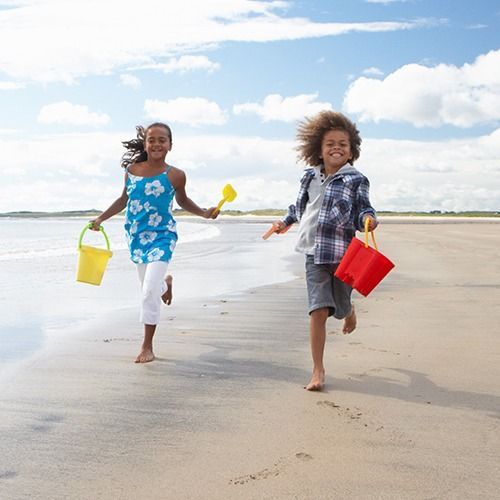 Two children running barefoot on a beach, holding colorful buckets under a bright sky.