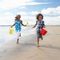 Two children running on a beach holding yellow and red buckets under a bright sky