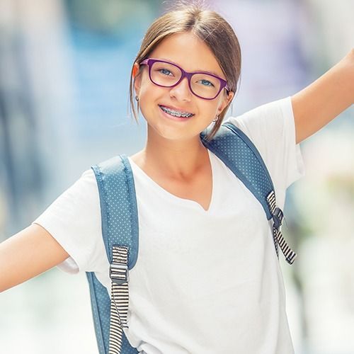 Smiling child with pink glasses and blue backpack, arms outstretched on a bright outdoor background