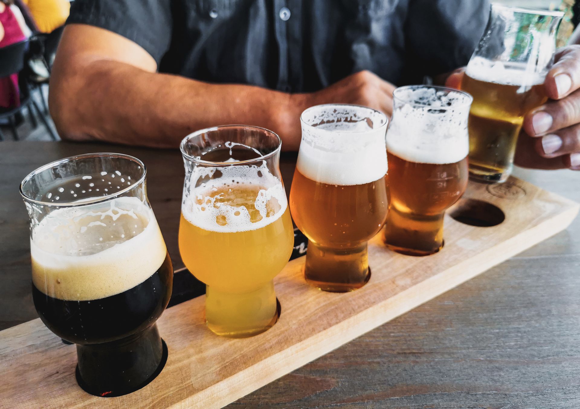 Flight of five beers on a wooden tray at a bar table, including a dark stout and pale ales