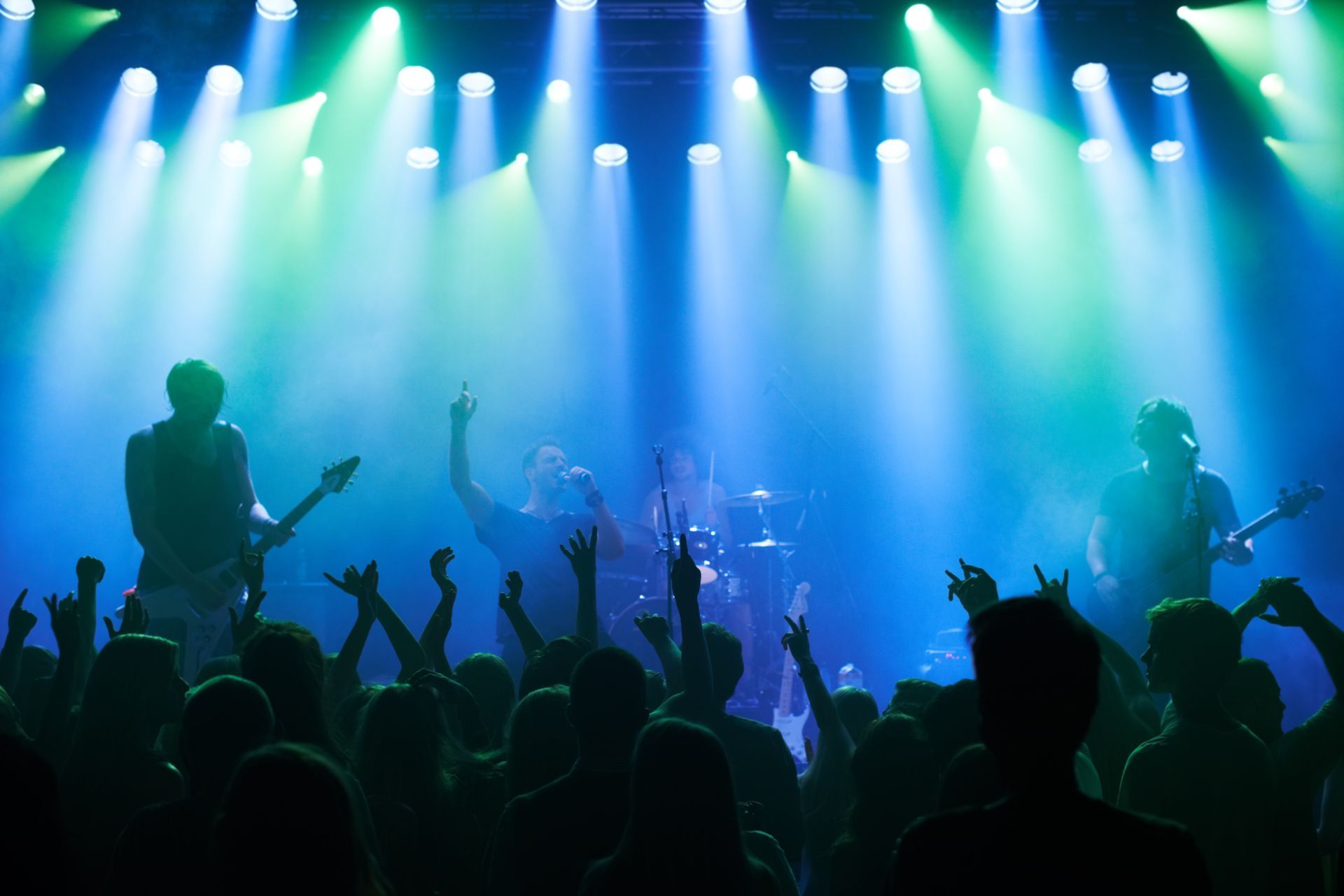 Silhouetted band performing on blue-green lit stage with crowd raising hands