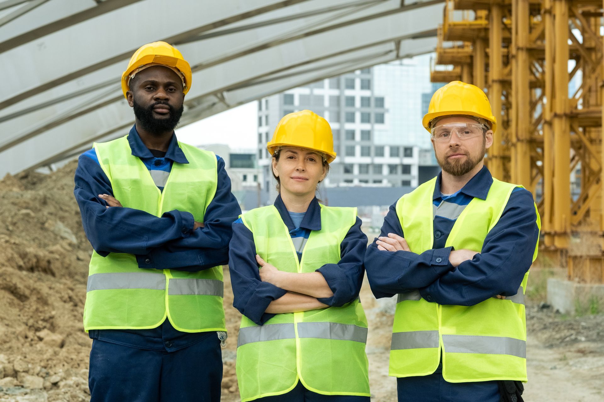 Three construction workers in yellow hard hats and green safety vests standing with arms crossed at a worksite